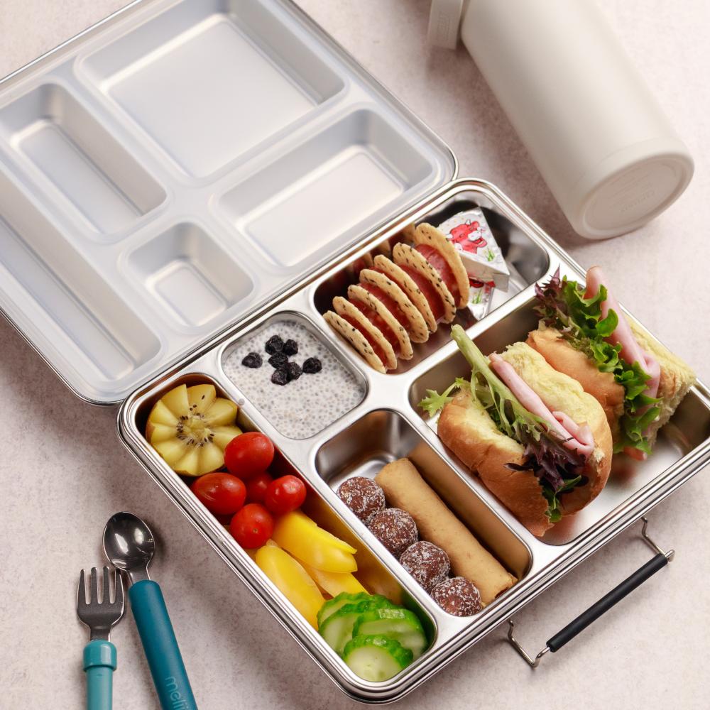 Metal lunch box with compartments filled with various food items on a light surface.