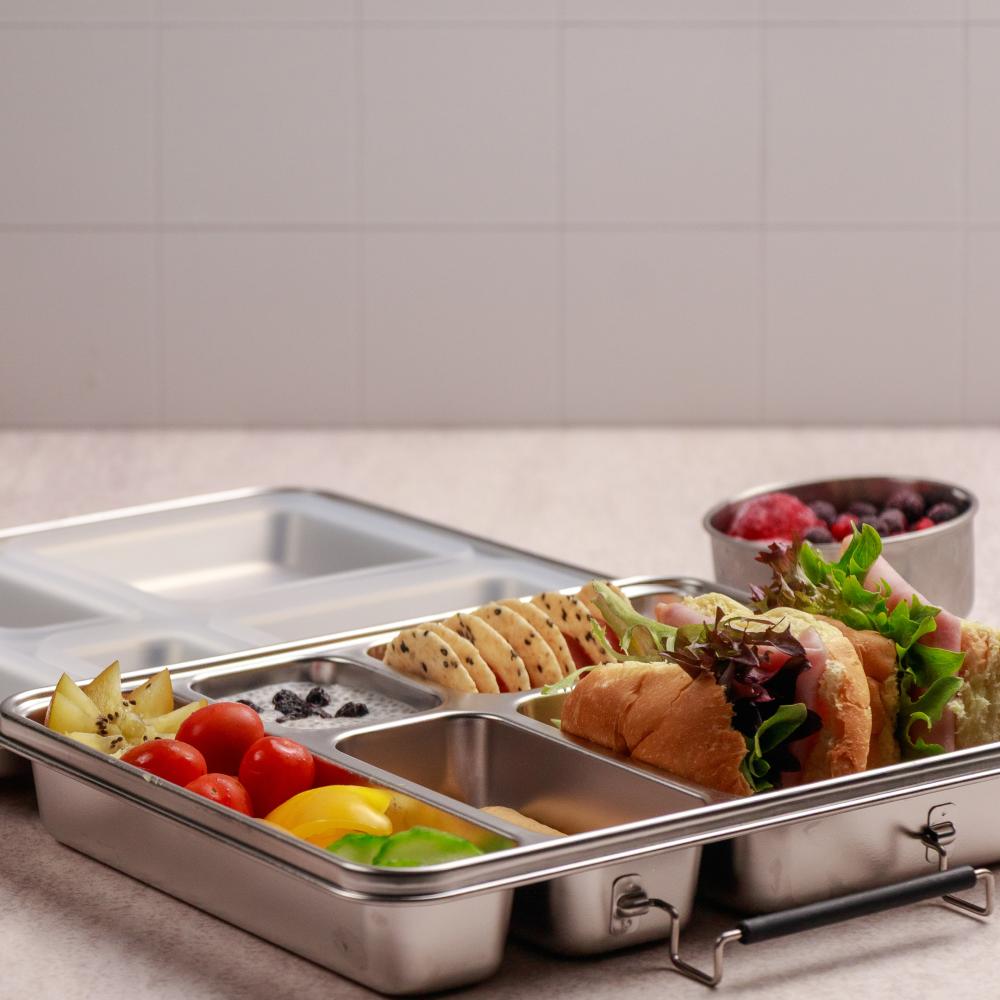 Stainless steel lunch box with compartments containing sandwiches and fruits on a white surface.