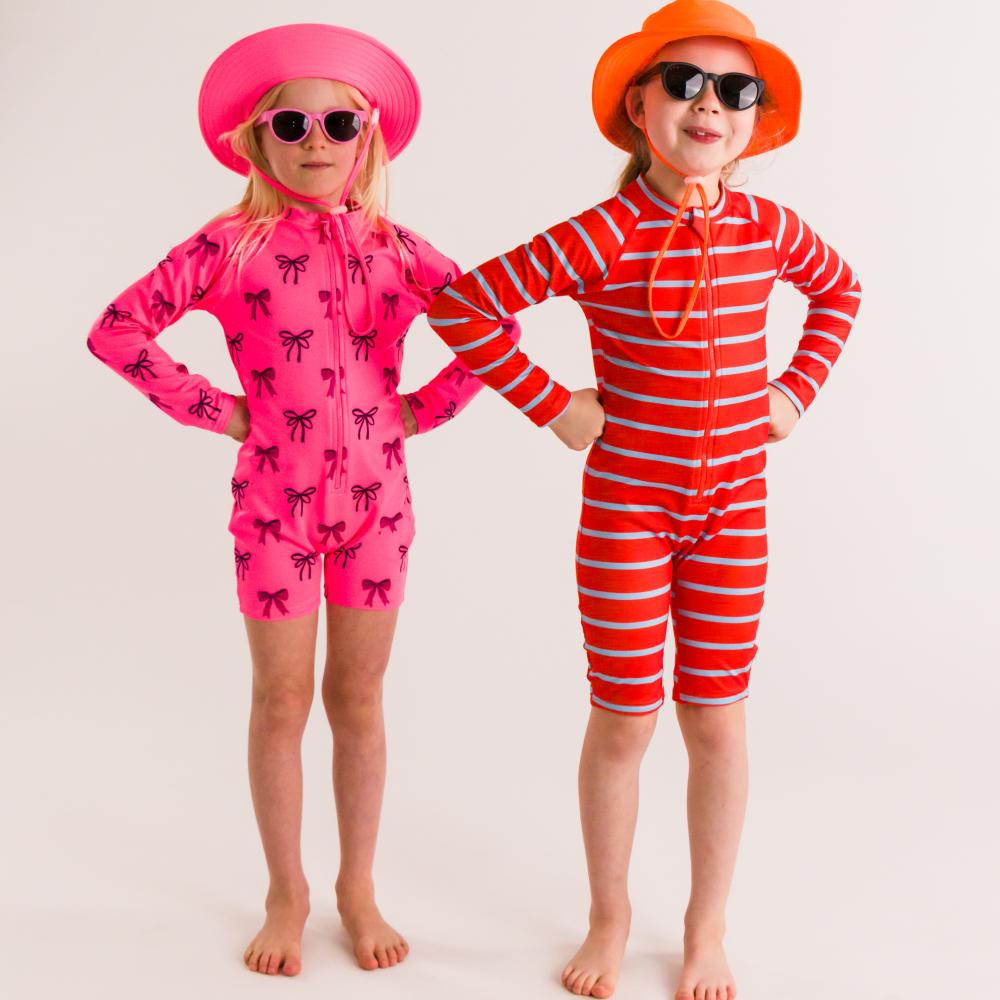 Two children wearing round ro.sham.bo sunglasses in colourful swimsuits and hats on a plain background