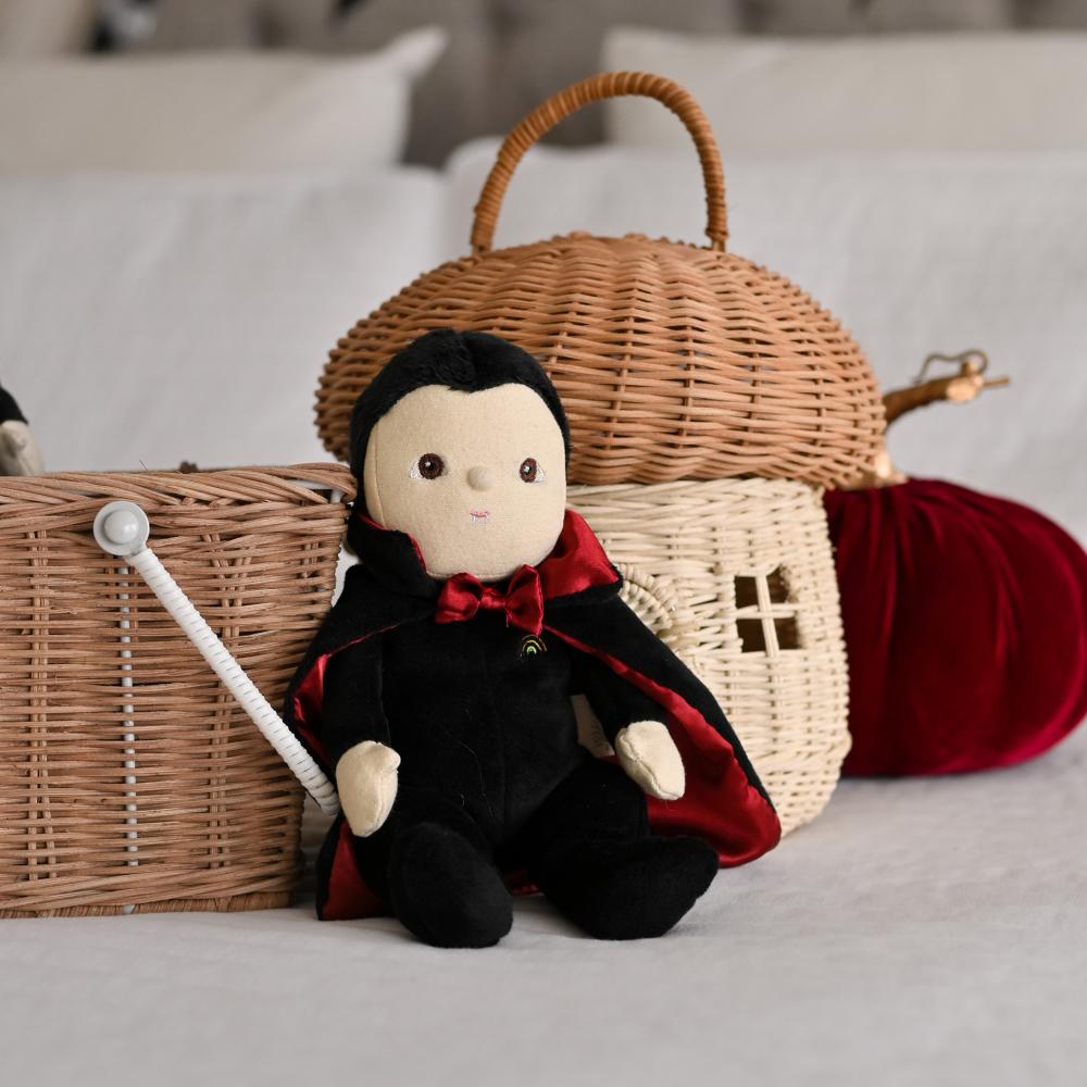 Stuffed toy in a black outfit with red cape next to a wicker basket on a light surface.