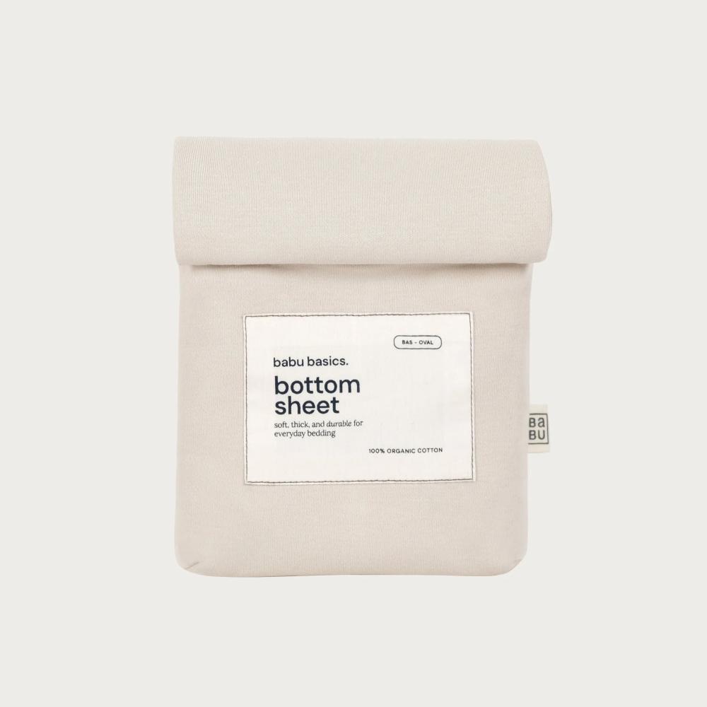 A folded light beige bottom sheet with a white paper label reading 'babu basics. bottom sheet' and a small side tag, shown on a plain light gray background.