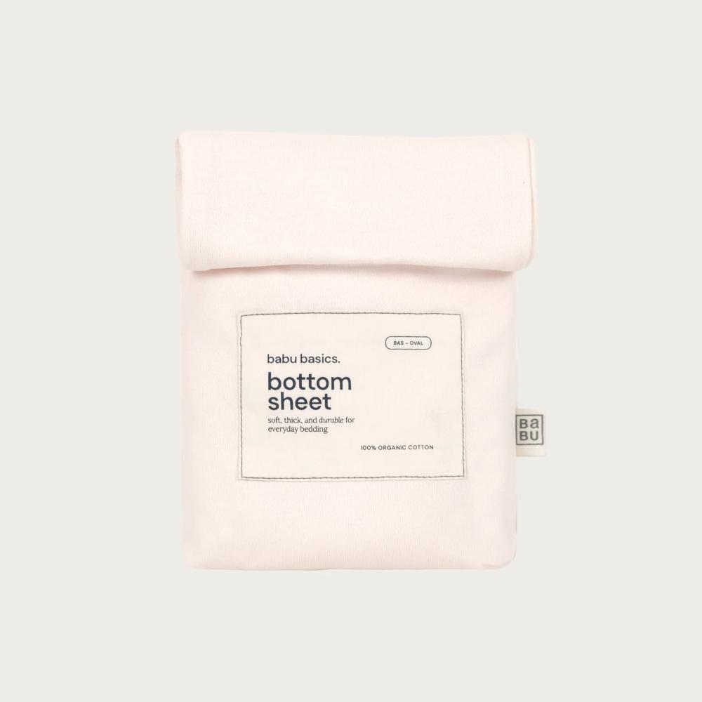 A pale pink folded cotton bottom sheet in its branded paper sleeve packaging, shown against a solid light gray background. The packaging features a white rectangular label with black text reading 'bubu basics.
