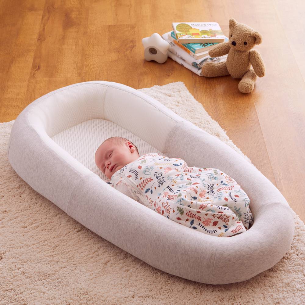 Baby sleeping in a soft grey mesh base baby pod on a wooden floor with a teddy bear and books in the background.