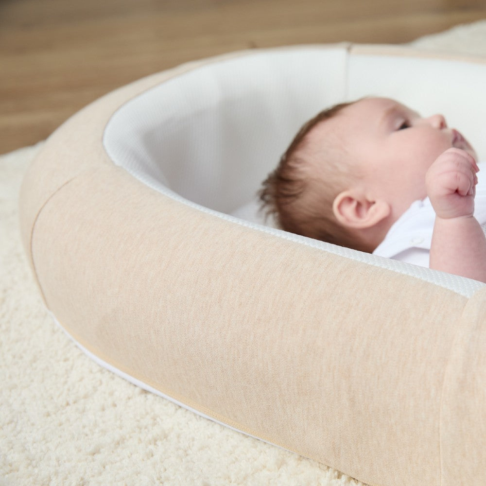 Baby lying on a beige and white baby nest with breathable mesh sides on a wooden floor.