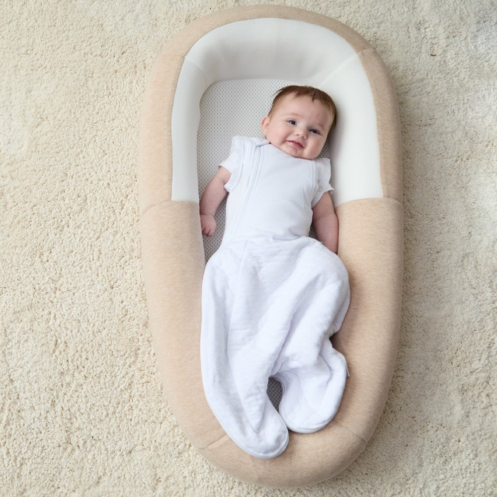 Baby lying in a beige and white baby nest on a carpeted floor