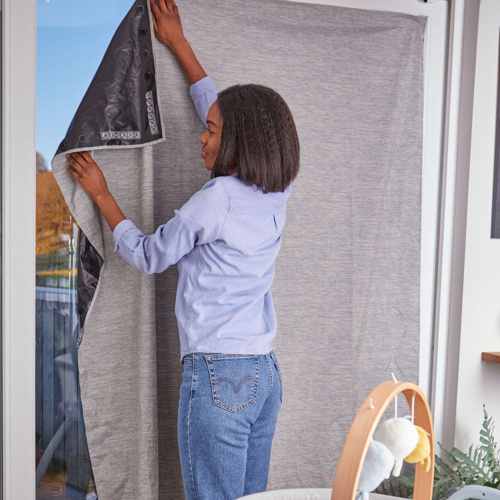Person installing a gray window blind in a room with a plant and lamp.