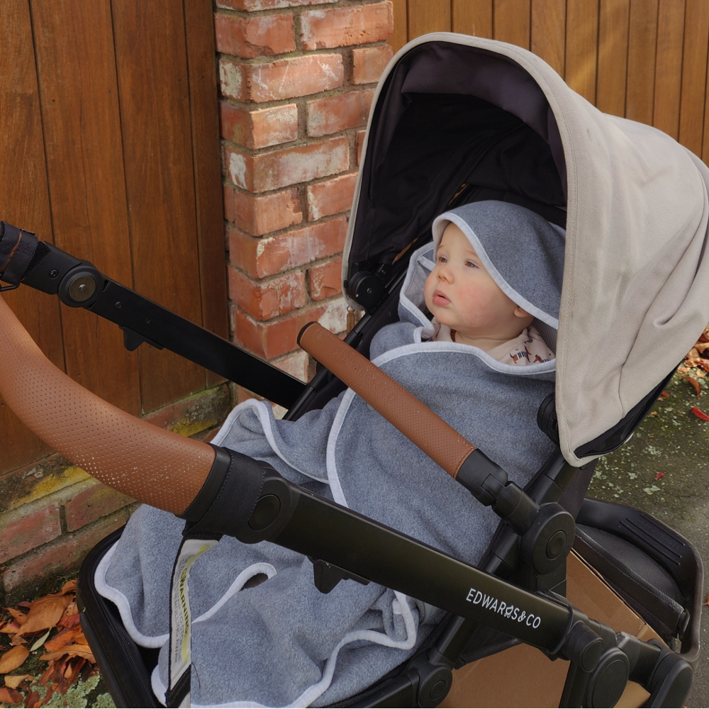 Child in a stroller with a hooded Purflo travel blanket, sitting outdoors next to a brick wall.