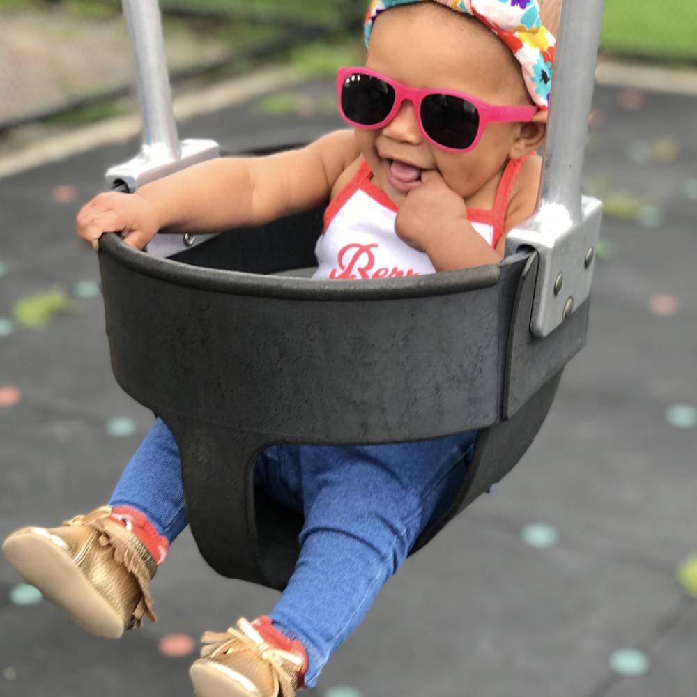 A baby sitting in a black playground swing seat outdoors, wearing bright pink sunglasses, a colorful patterned headband, a white tank top with red trim, blue jeans, and gold moccasin-style shoes.