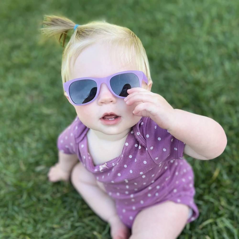 A baby wearing lavender sunglasses with dark lenses and a matching purple polka-dot romper, sitting on green grass outdoors.