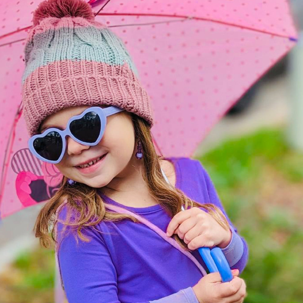 Child wearing a pink knit hat and heart-shaped sunglasses, holding a pink umbrella 