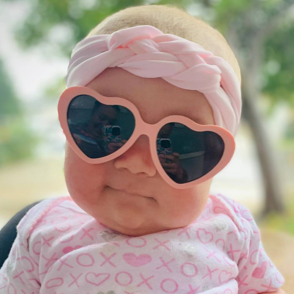 A close-up of a baby wearing heart-shaped pink sunglasses, a pink braided headband, and a pink patterned top, photographed outdoors with a blurred natural background.