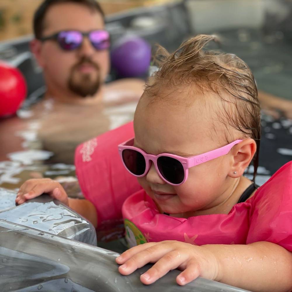 Child wearing pink sunglasses and a life jacket in a spa pool with a person in the background.