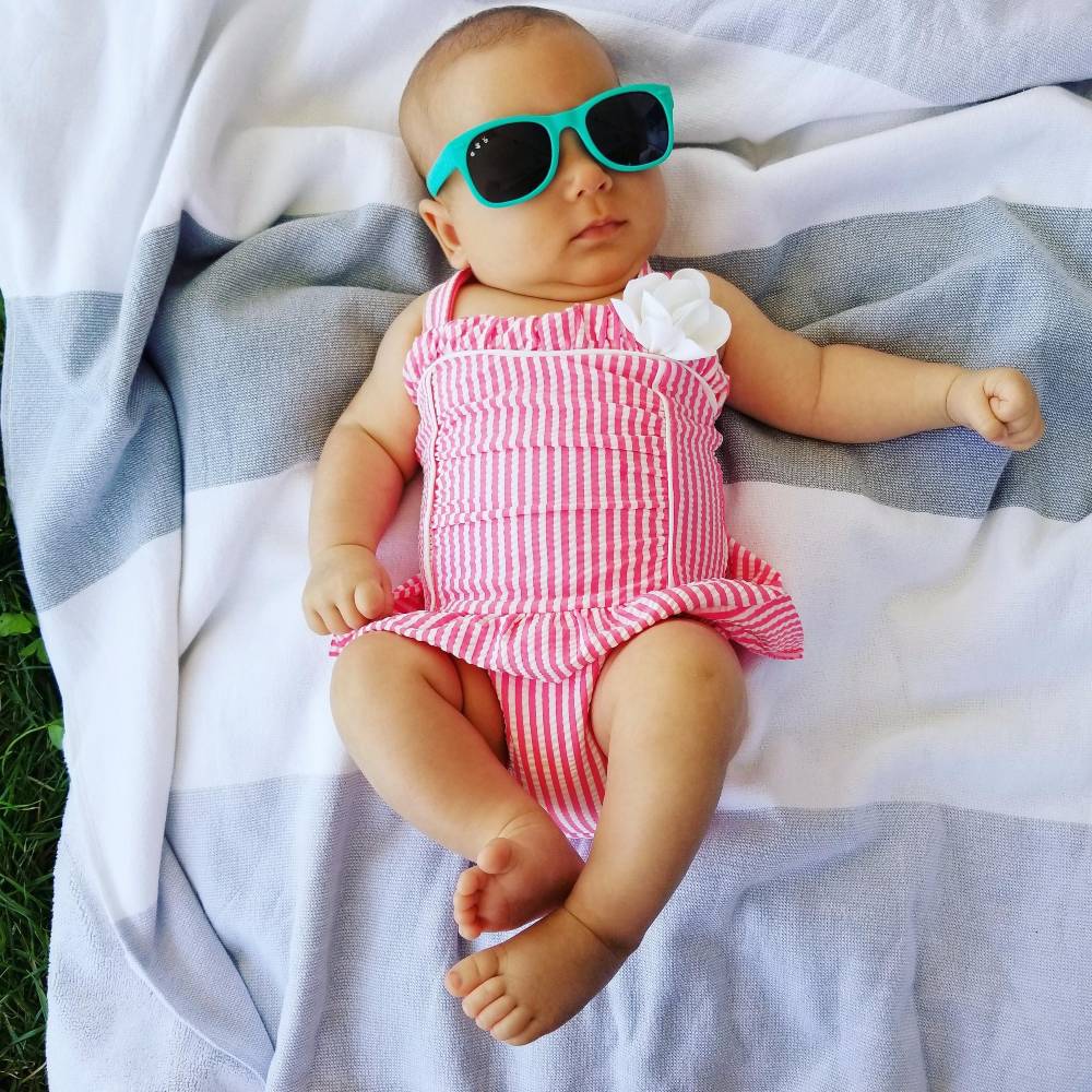 A baby lying on a striped blanket, wearing a pink and white striped romper with a white flower detail and bright green sunglasses.