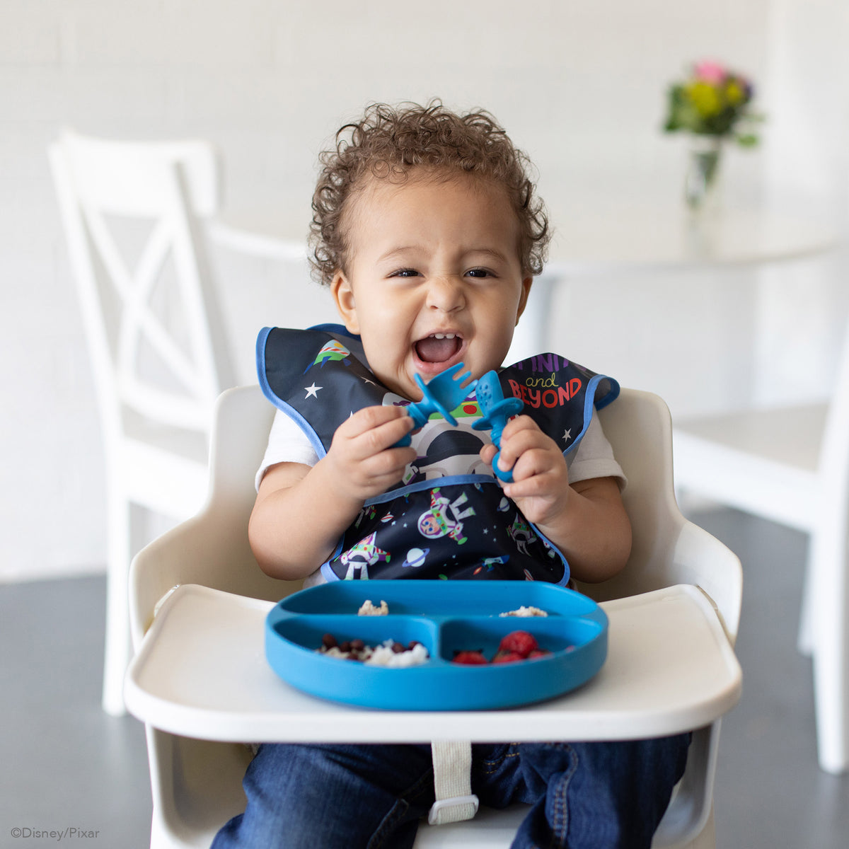 Blue Silicone Grip Dish with food inside on a white highchair tray with a curly haired child in a navy blue bib holding dark blue chewtensil cutlery 