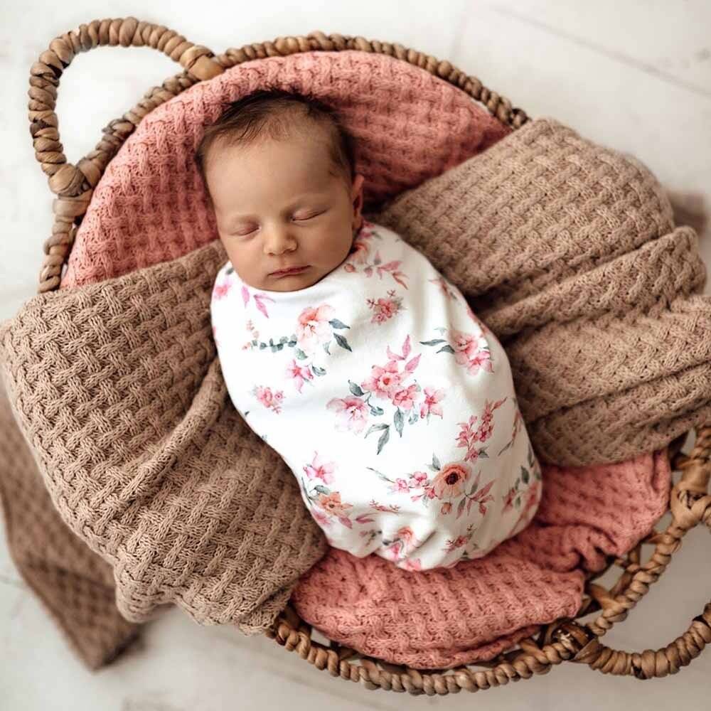 A baby lying in a basket, swaddled in a Snuggle Swaddle with a floral design.