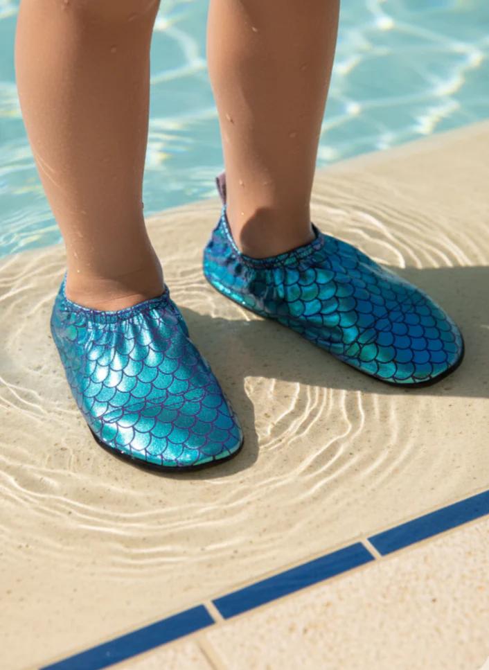 Blue/green mermaid scale-patterned shoes worn by a person standing in pool near water.