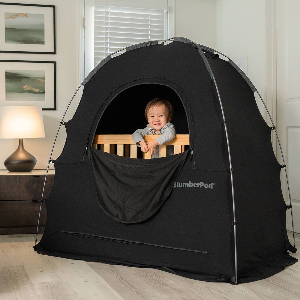 Child peeking out from a black SlumberPod in a room with framed pictures on the wall.