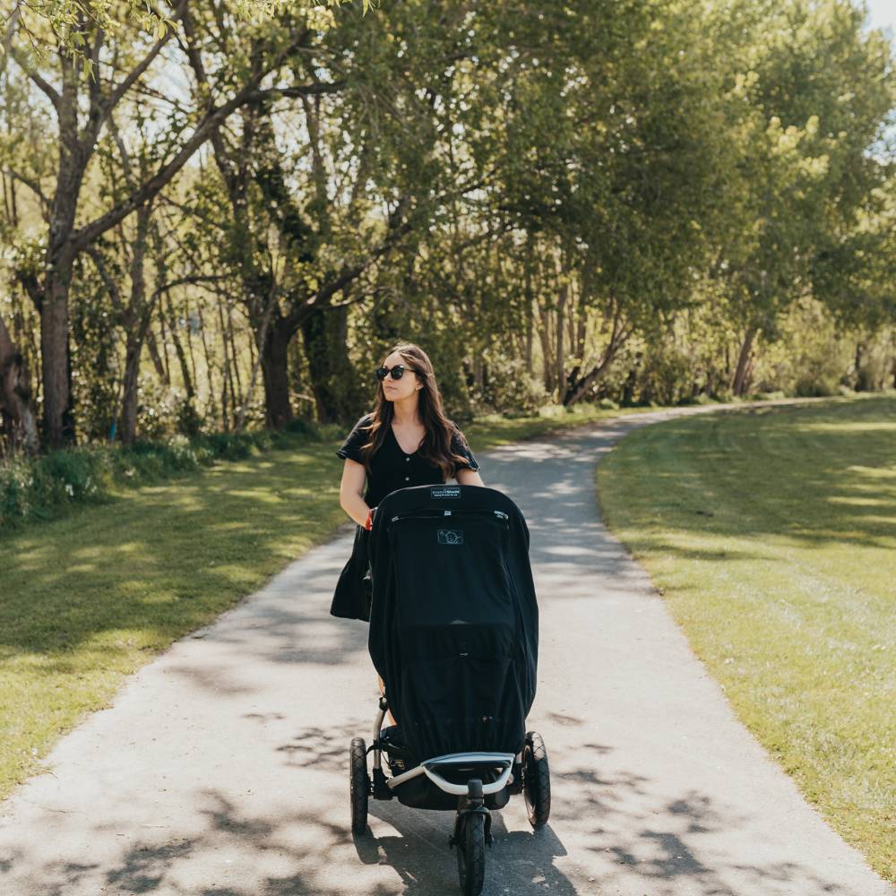 Woman pushing a stroller with Snoozeshade Blackout cover on a path in a park with trees and grass.