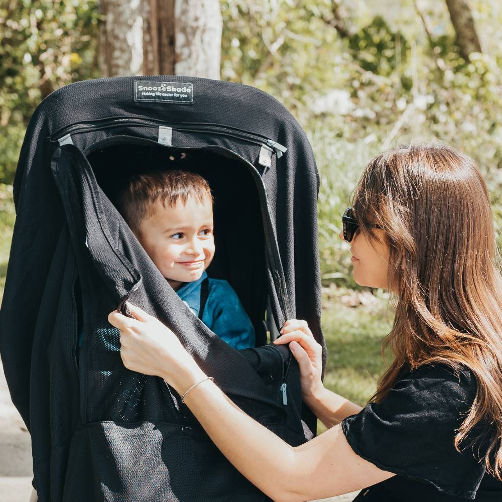 Woman adjusting a Snoozeshade stroller cover for a child outdoors.