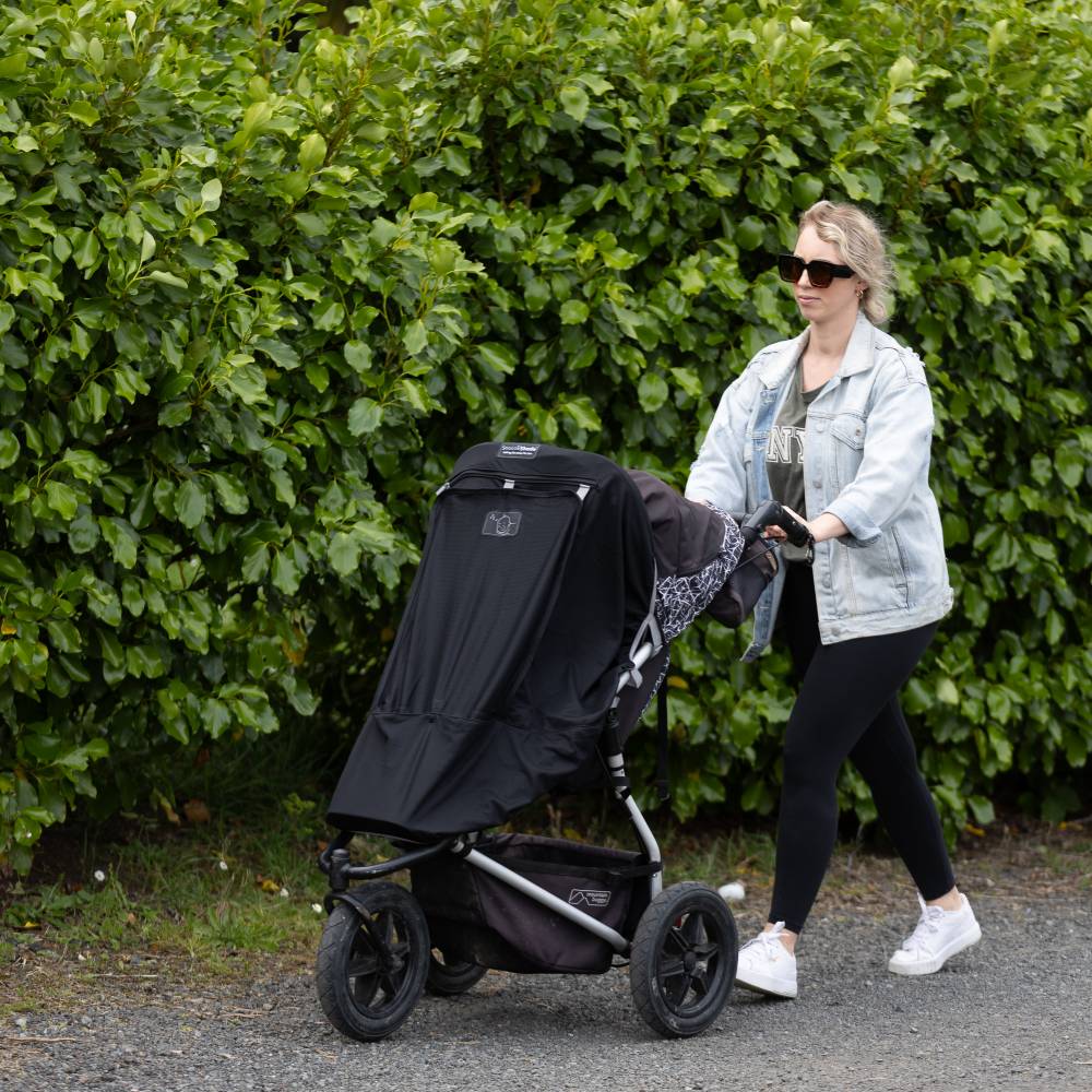 Woman pushing a stroller with Snoozeshade blackout cover against a green hedge.