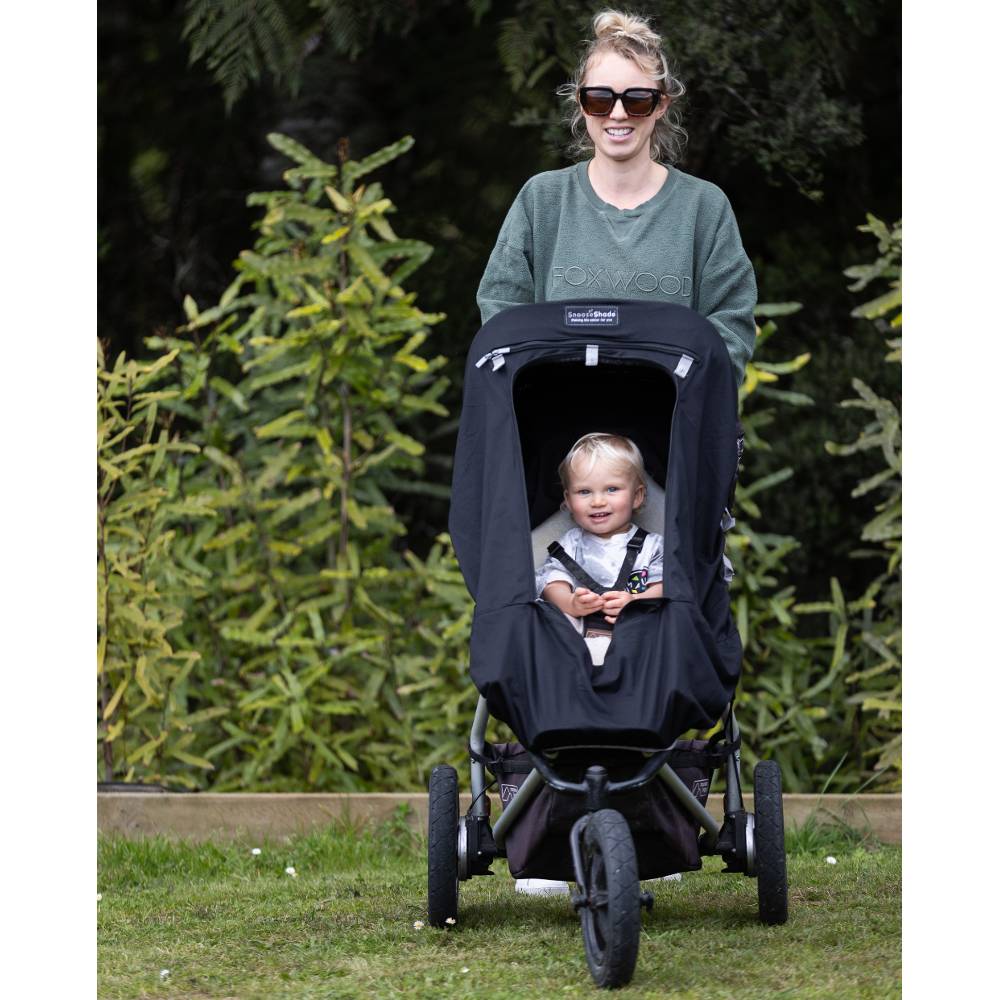 Woman pushing a stroller with Snoozeshade blackout cover with a child inside, standing outdoors with greenery in the background.