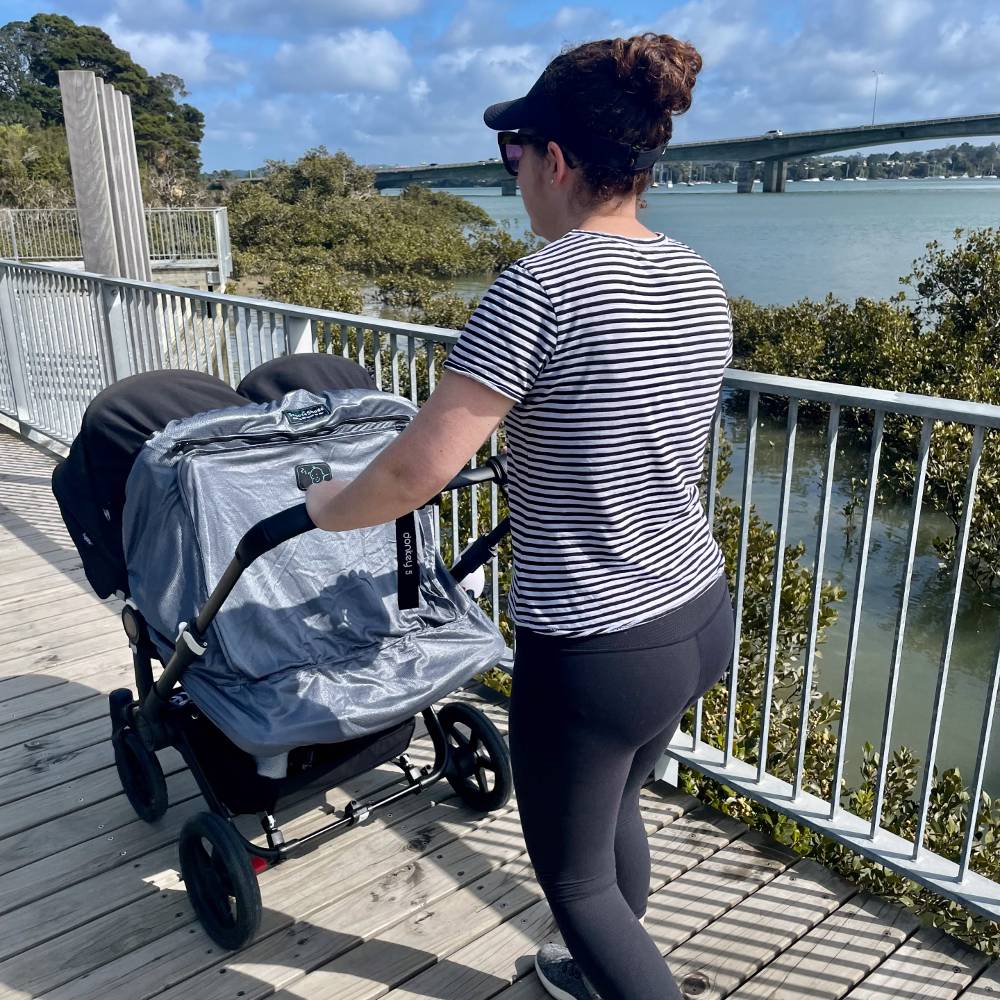 Woman pushing a twin stroller with a Snoozeshade blackout cover by a waterfront with a bridge in the background