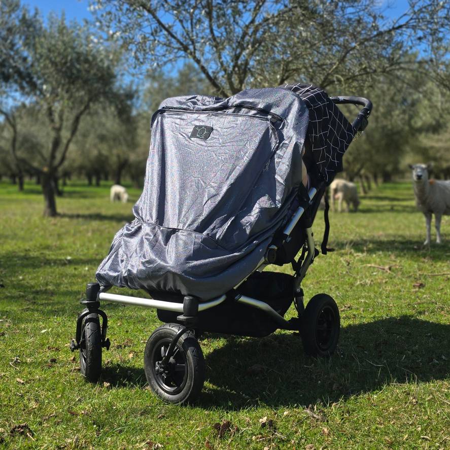 Twin stroller with a blackout cover in a grassy field with sheep in the background