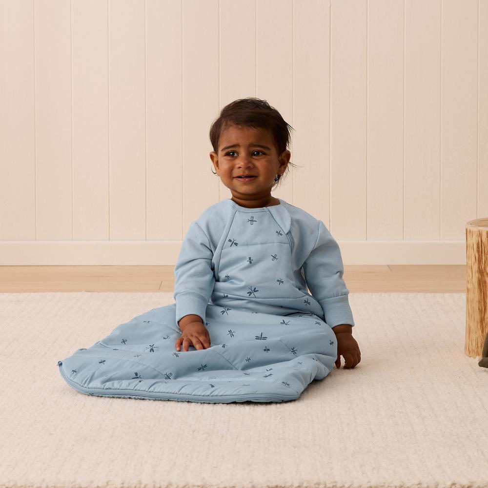 Child wearing a light blue sleep sack sitting on a beige floor.