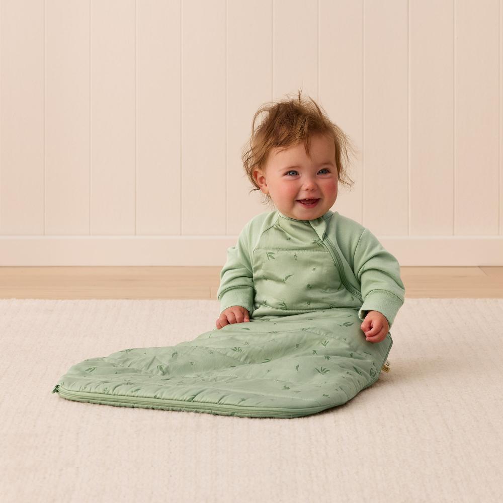 Baby in a green sleeping bag sitting on a light wooden floor.