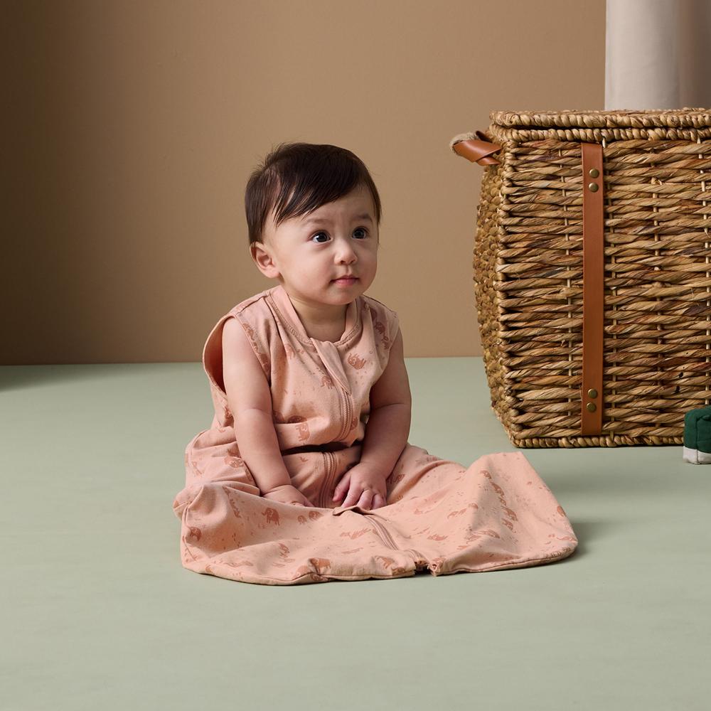 Baby in a pink sleep sack sitting on a green floor with a woven basket in the background.