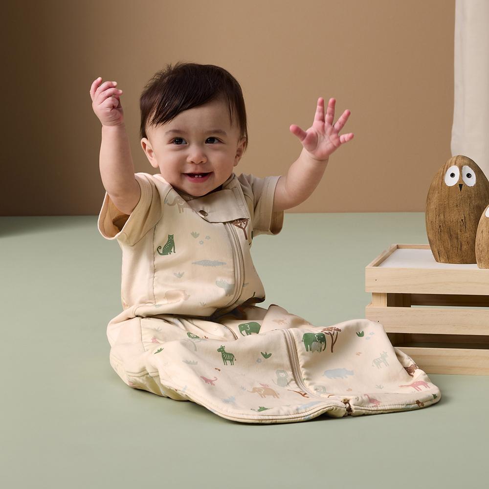 Baby sitting on a green floor with arms raised, wearing a beige outfit with animal prints.