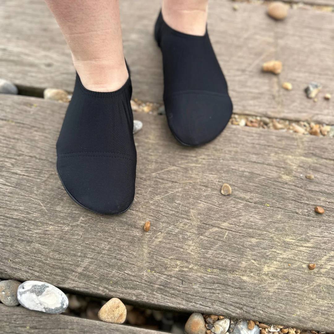 Black slipfree water shoes worn on a wooden deck with pebbles.