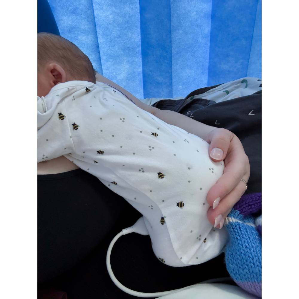 Baby wearing a white onesie with bee pattern, held by a person against a blue curtain background.