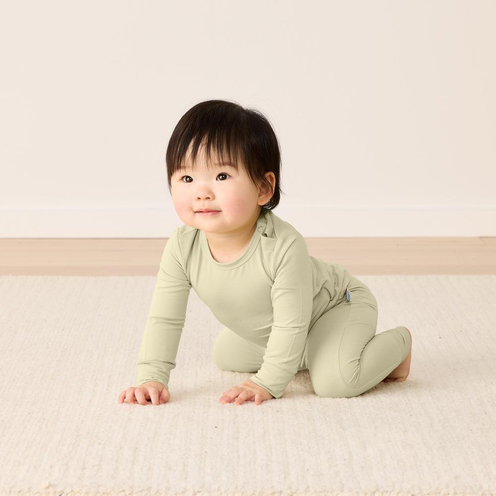 Baby in a green outfit sitting on a light-colored floor.