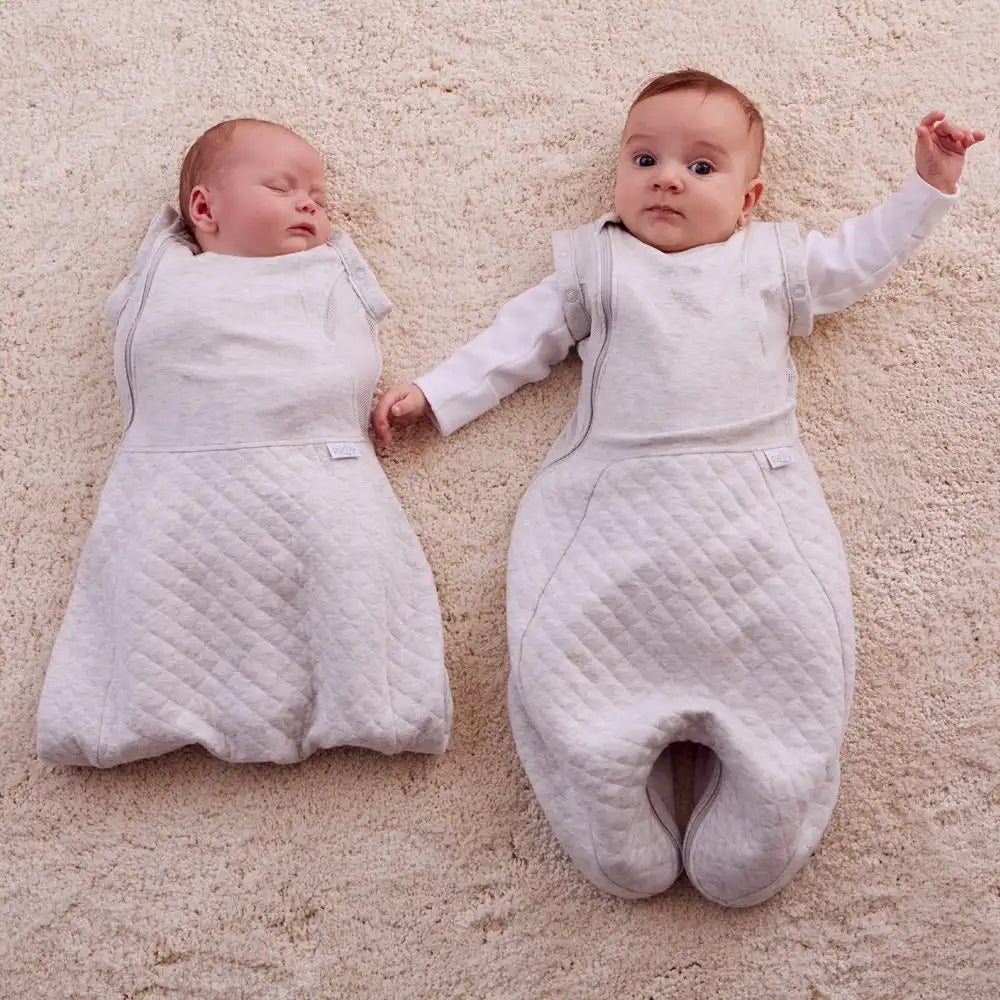 Two babies wearing gray sleep sacks on a beige carpet with arms in and out