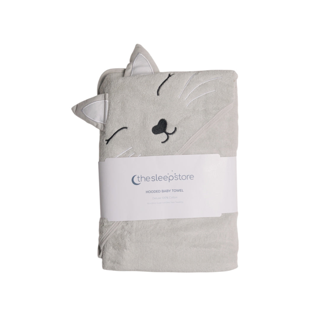 A folded light gray terry cotton hooded baby towel shaped like a cat face, with embroidered closed eyes, nose, and ears, wrapped in a white paper band labeled 'thesleepstore'.