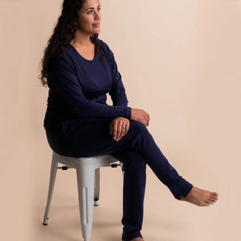 A woman with long curly dark hair sits on a white metal stool, wearing a matching navy blue long-sleeve top and pants set against a solid beige background.