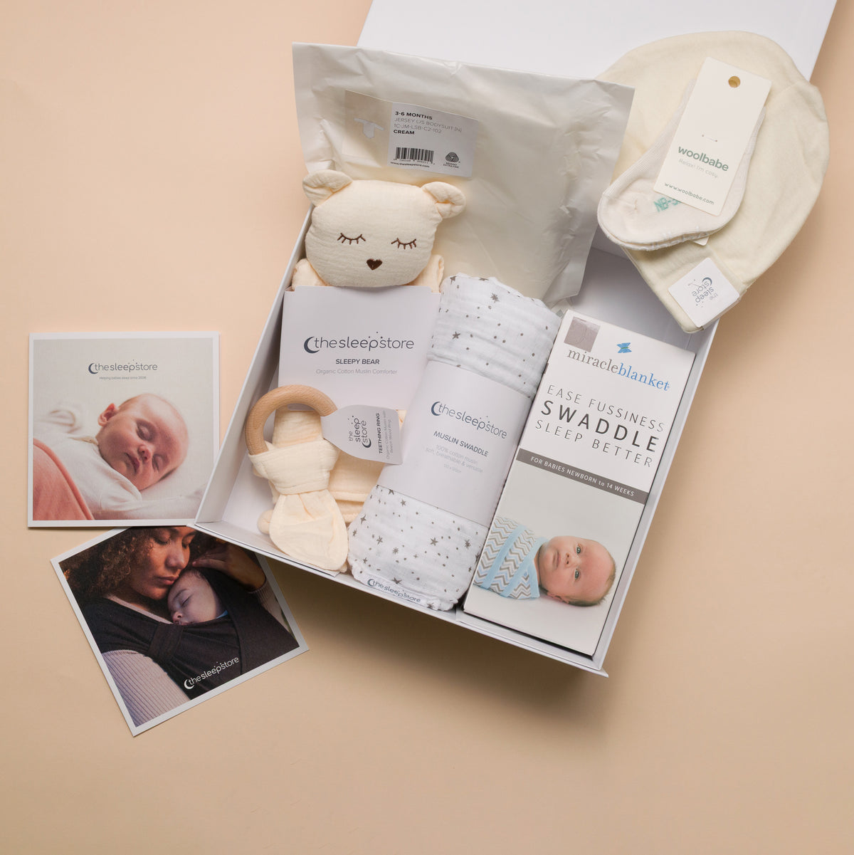 A baby gift box set from the Sleepstore, featuring a plush bear toy, swaddles, a wooden teether, a muslin blanket, and care instructions, arranged on a beige background with two printed photos.