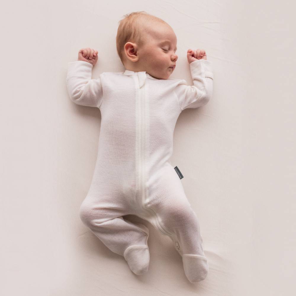 Baby lying on a white surface wearing a white jersey merino footed onesie.