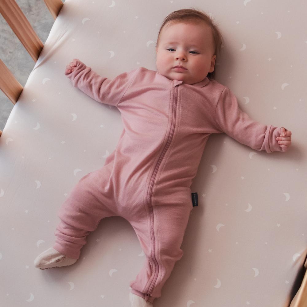 Baby in a pink merino onesie lying on a crib with a polka dot mattress cover.