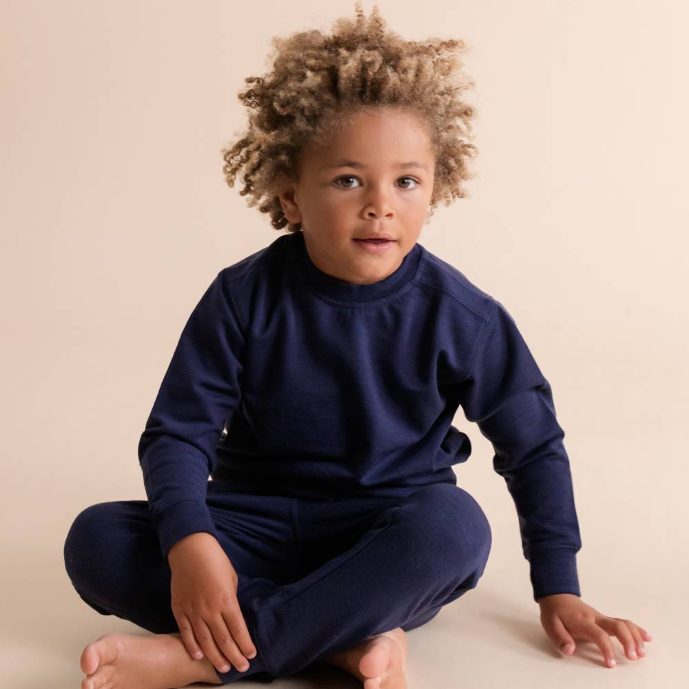 A young child with curly light brown hair sits cross-legged on a plain beige studio background, wearing a matching navy blue long-sleeve top and merino fleece jogger pants set.