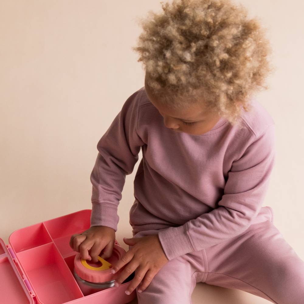 A young child with curly light brown hair sits on a beige floor, wearing a dusty mauve pink merino fleece sweatshirt and matching pants. They are interacting with a pink divided lunch box containing a small pink and yellow container.