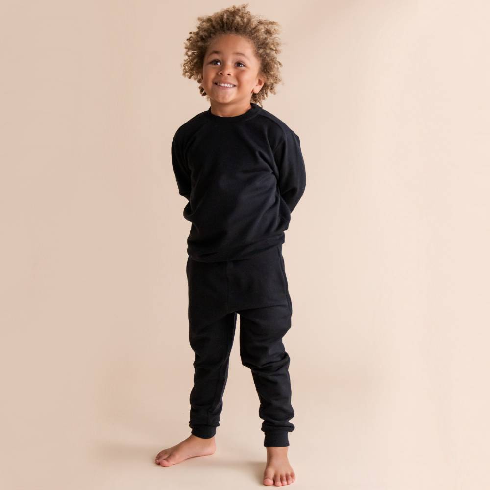 A young child with curly hair stands barefoot against a solid beige studio background, wearing a matching black long-sleeve sweater and black jogger-style pants in a merino fleece fabric