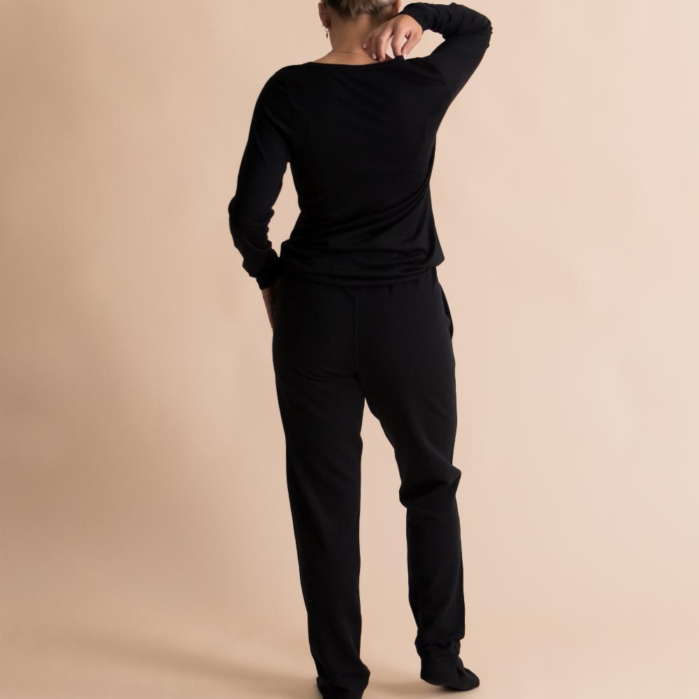 A woman wearing a black long-sleeve top and black pants, shown from the back against a solid beige studio background.