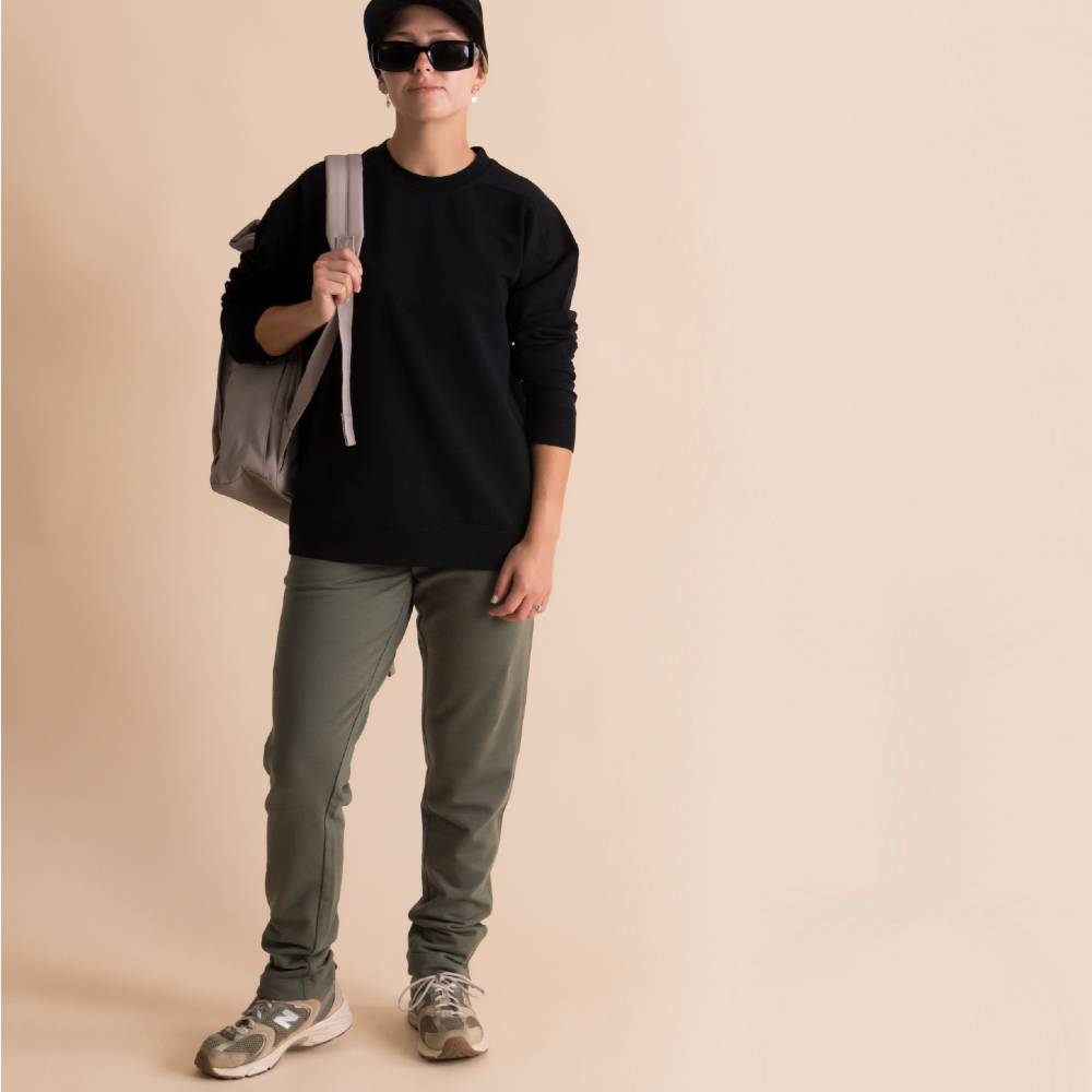A person modeling a black long-sleeve merino fleece crewneck sweatshirt, olive green pants, and gray sneakers, carrying a gray tote bag over their shoulder, set against a solid beige background.