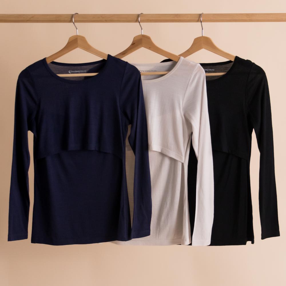 Three long-sleeve Merino Tencel Breastfeeding shirts in navy, white, and black hanging on a rack against a beige background.