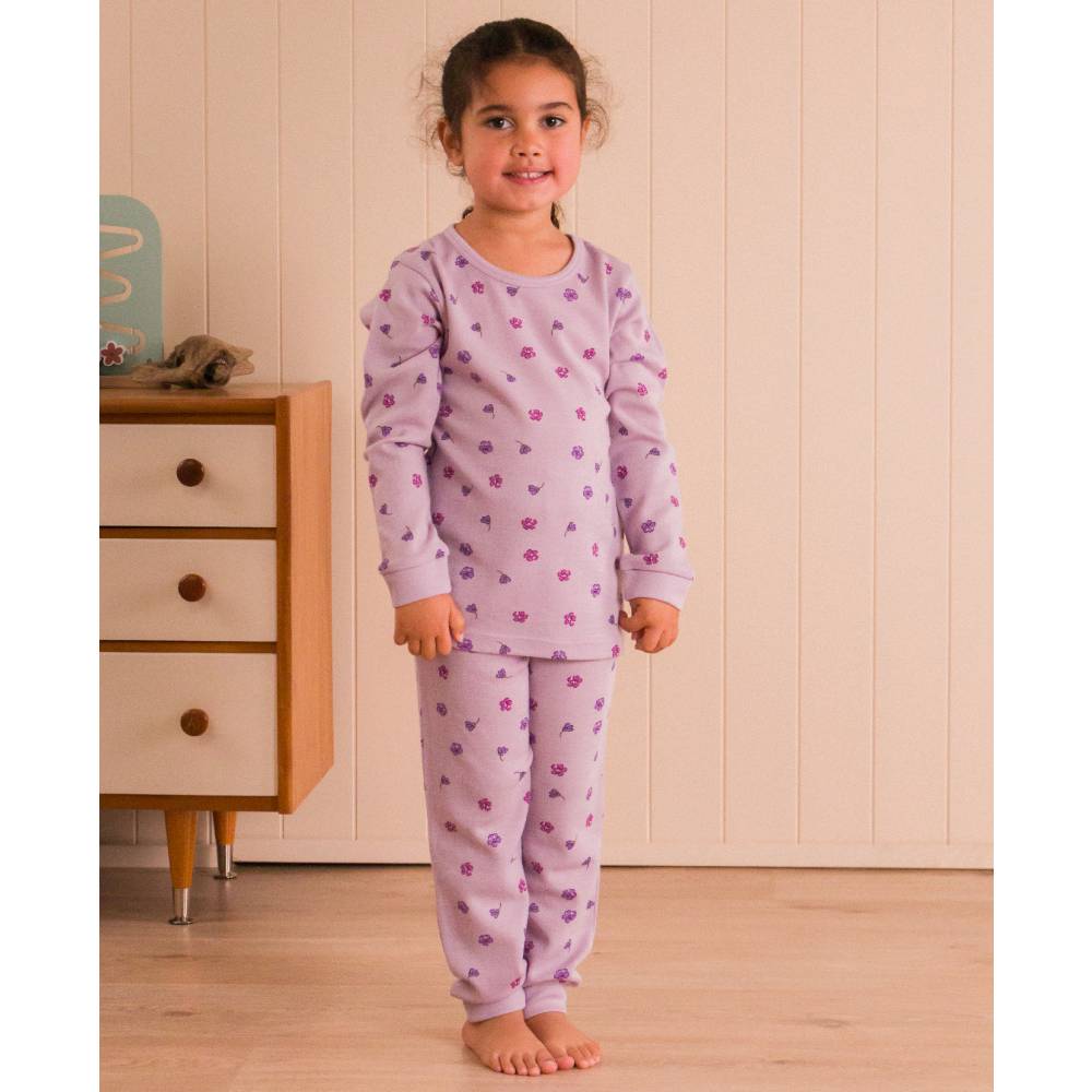 Child wearing a purple pajama set with a floral pattern in a room with wooden flooring and a dresser.