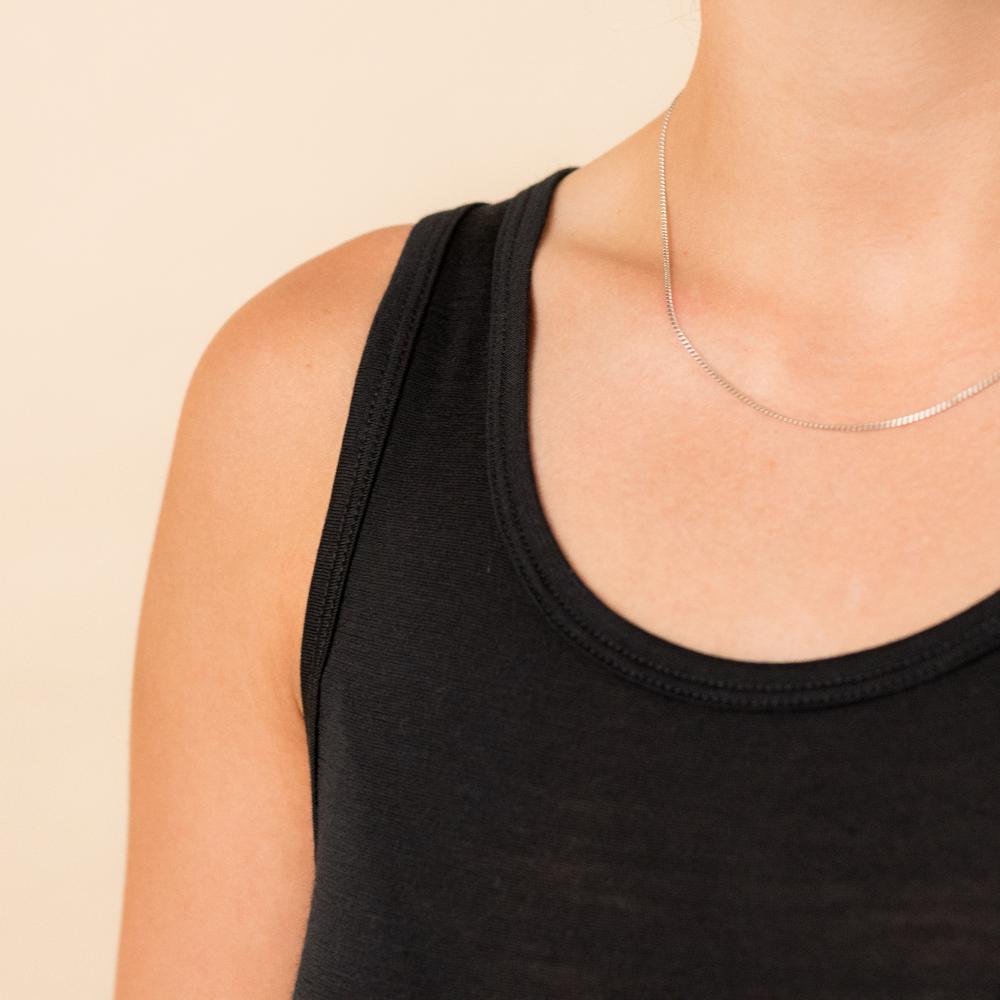 Close-up of a person wearing a thin silver chain necklace with a black ribbed tank top against a plain beige background.