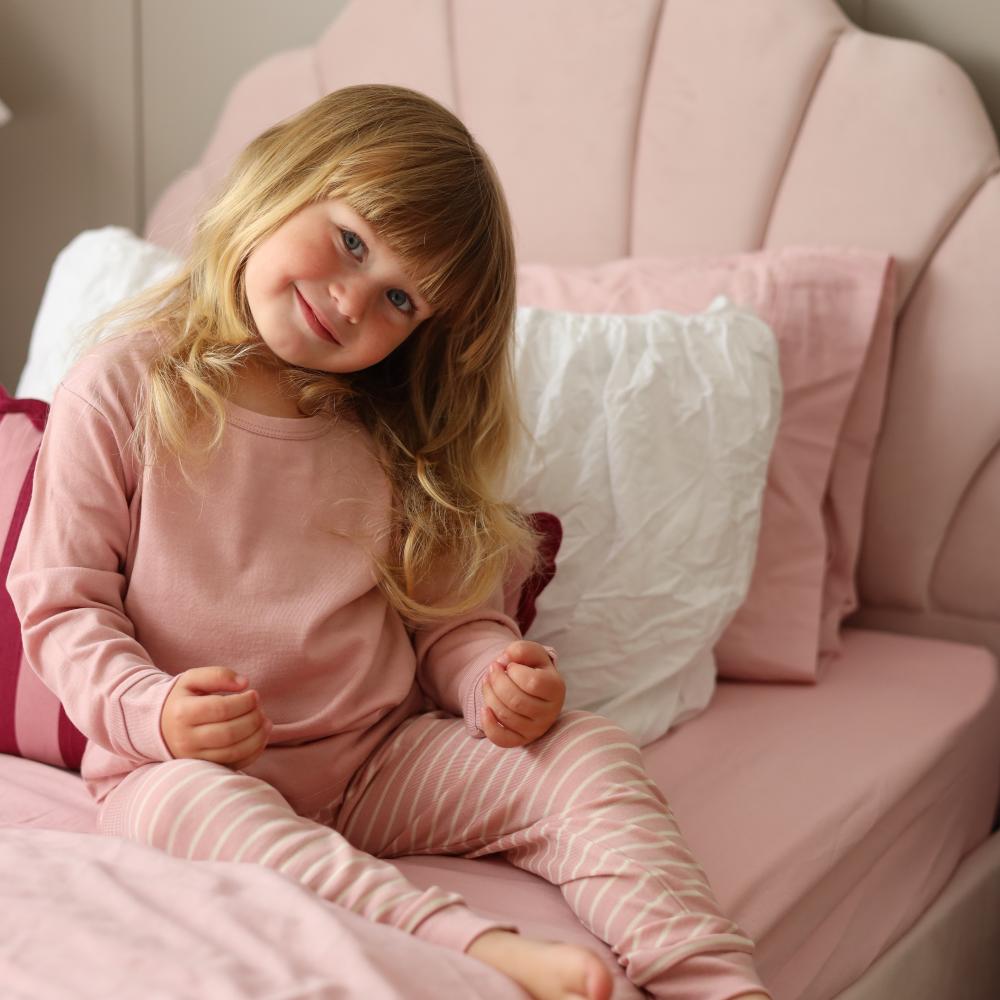 A young girl with blonde hair sits on a pink bed wearing a matching dusty pink jersey merino long-sleeve top and pink-and-white striped pajama pants.