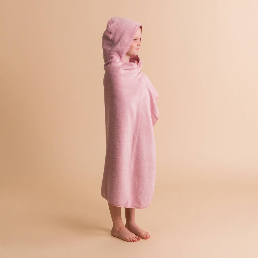 A child standing in profile wearing a soft pink hooded towel against a plain warm beige studio background.